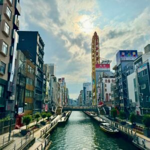 A bustling urban canal scene, lined with tall buildings and billboards. A ferris wheel towers above, under a cloudy sky with the sun peeking through.