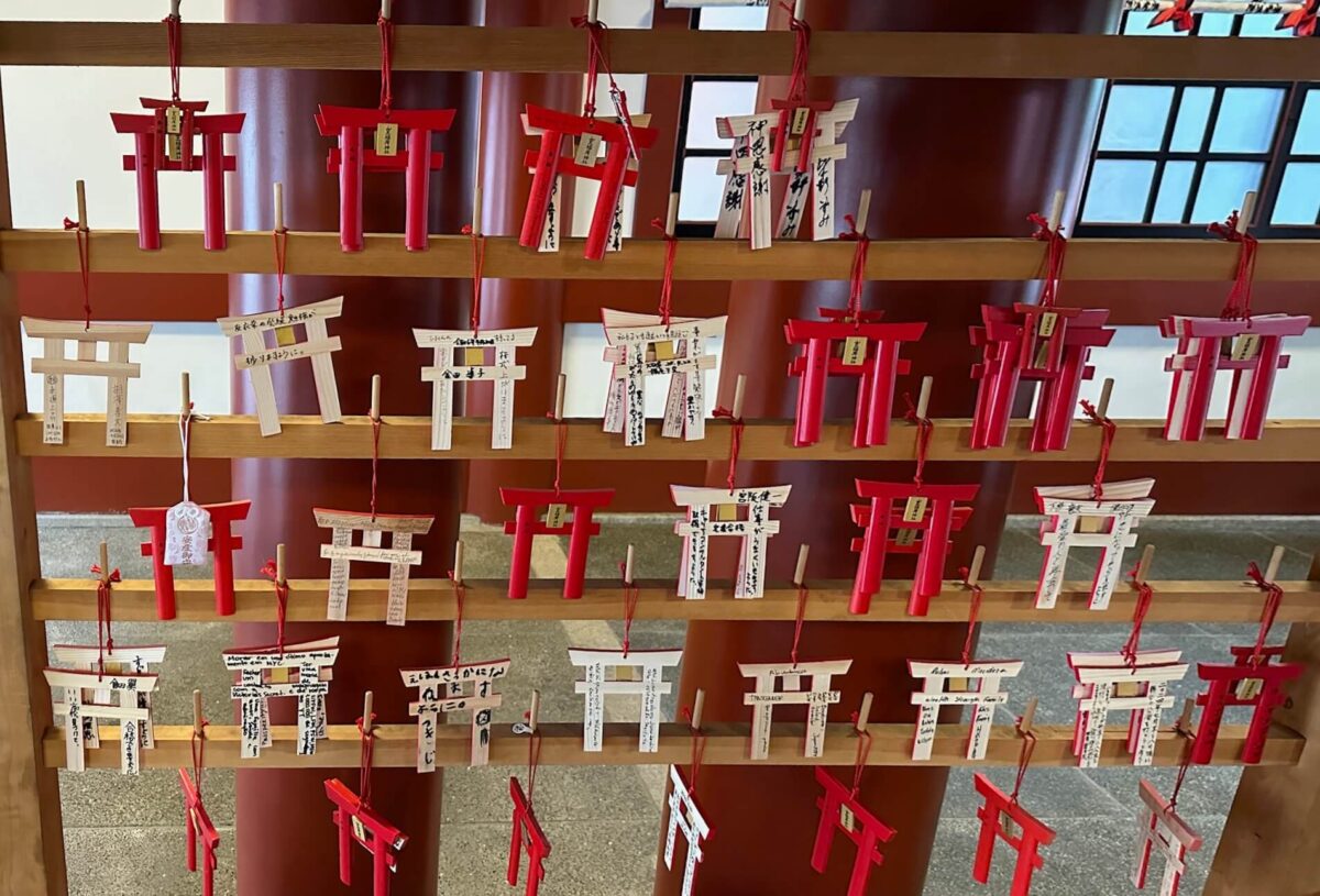 Racks display small red and white wooden torii gates with handwritten messages hanging by red strings, creating a peaceful, reflective atmosphere.
