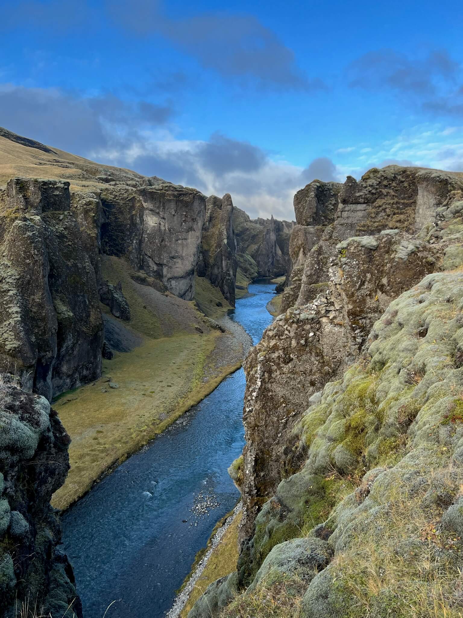 A serene landscape of a deep canyon with a winding river, framed by mossy cliffs. The sky is blue with scattered clouds, conveying tranquility.