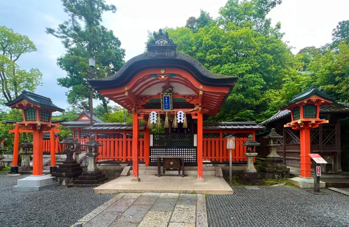 A traditional Japanese shrine with a red wooden structure and lanterns, surrounded by lush green trees. The atmosphere is peaceful and serene.