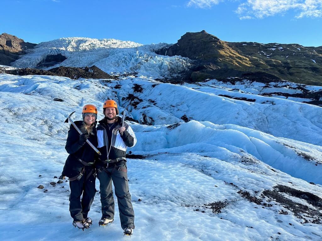 A smiling couple stands on a glacier, wearing helmets and holding ice axes. The bright blue sky and rugged ice formations create an adventurous tone.