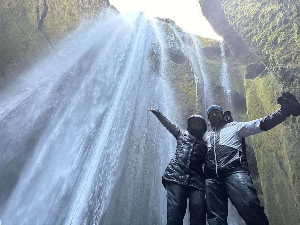 Two people in raincoats joyfully stand in front of a cascading waterfall in a mossy canyon, arms outstretched, conveying excitement and adventure.
