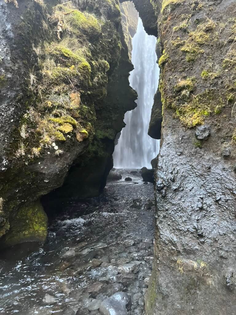 Narrow rocky gorge with lush moss-covered walls framing a distant waterfall cascading into a clear stream. The scene conveys tranquility and natural beauty.