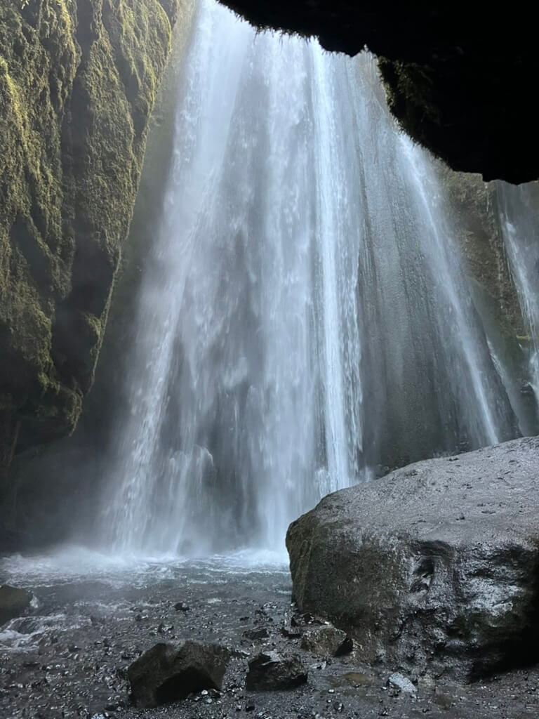 Waterfall cascading through a moss-covered cliff into a rocky pool below, creating a misty atmosphere. Sunlight filters in, adding a serene, majestic feel.