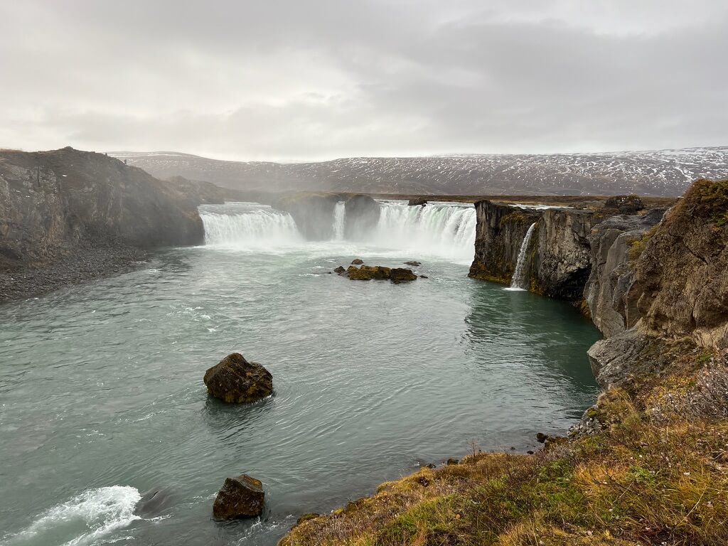 Majestic waterfall cascades over rocky cliffs into a wide river, creating mist. Overcast sky and rugged landscape evoke a serene, tranquil atmosphere.