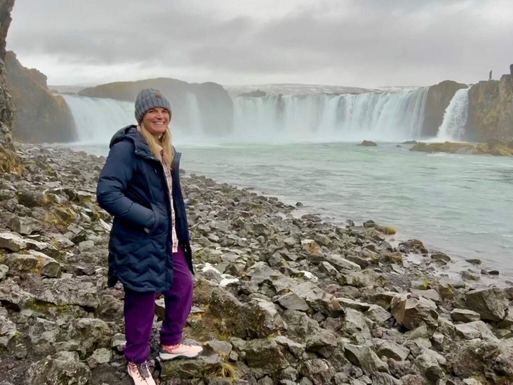 A woman in a black coat and gray beanie stands on a rocky shore, smiling in front of a wide waterfall under a cloudy sky, conveying a sense of adventure.
