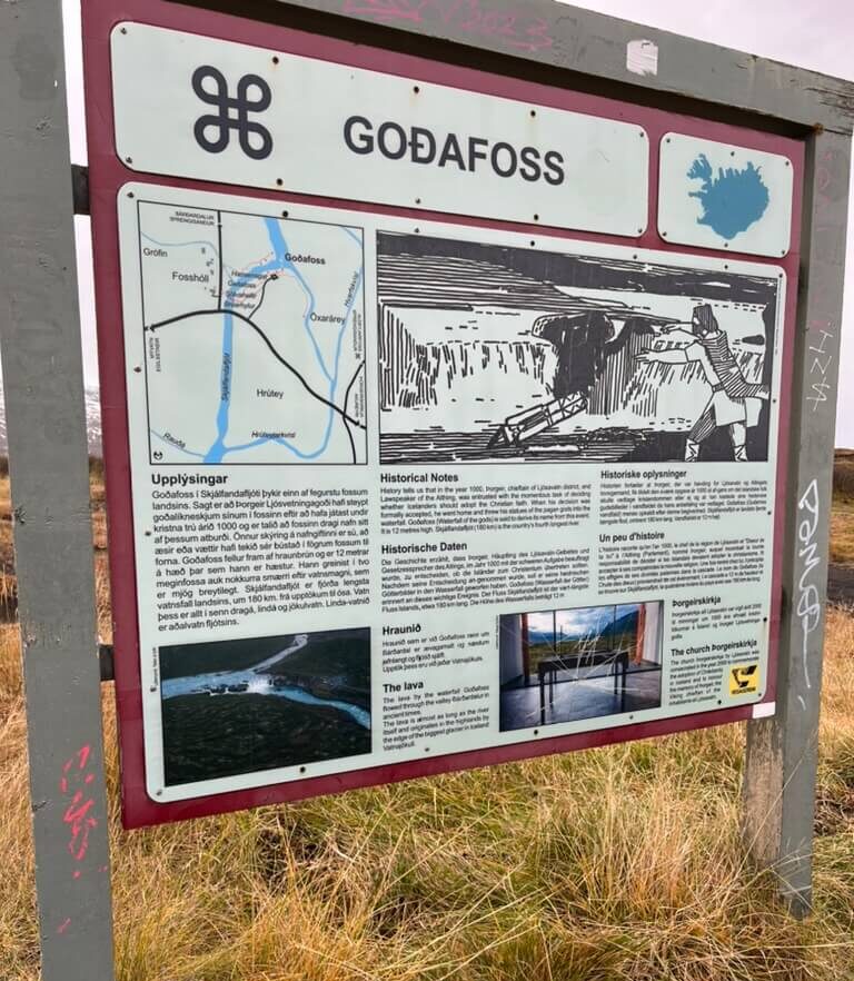 Informational sign for Goðafoss waterfalls with a map, historical and geological notes in multiple languages. Surrounded by grass and graffiti.
