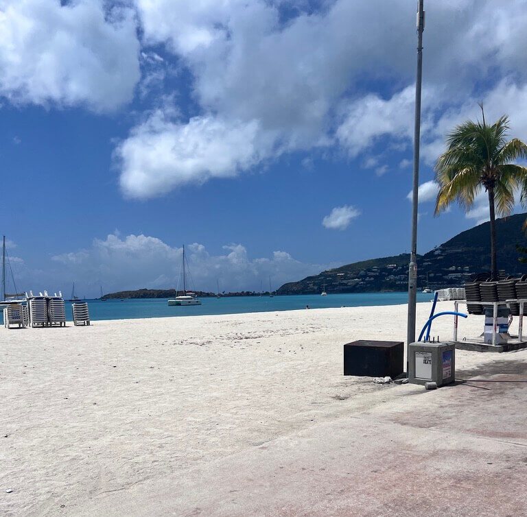 Sunny beach scene with a palm tree and stacked beach chairs on white sand. Calm blue sea with sailboats in the distance. Hills under a blue sky with clouds.