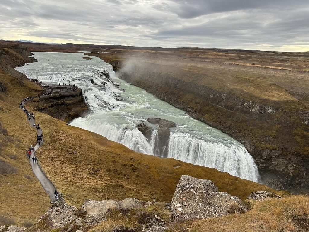 Winding path leads to a majestic waterfall cascading into a rugged canyon under overcast sky; visitors walk along the trail, capturing the serene landscape.