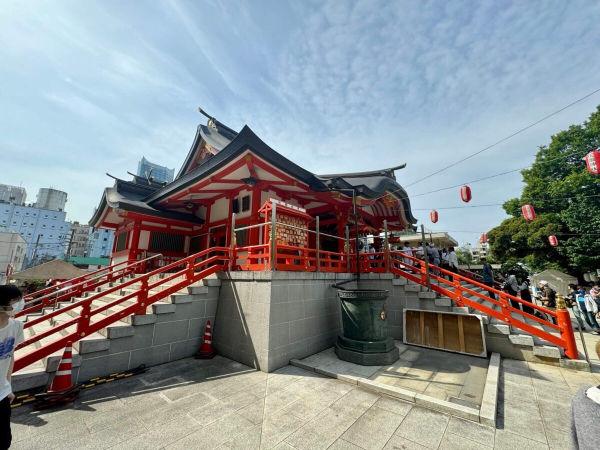 Traditional Japanese shrine with red railings and a gray stone base. Bright lanterns hang overhead; people gather outside. Sky is partly cloudy.