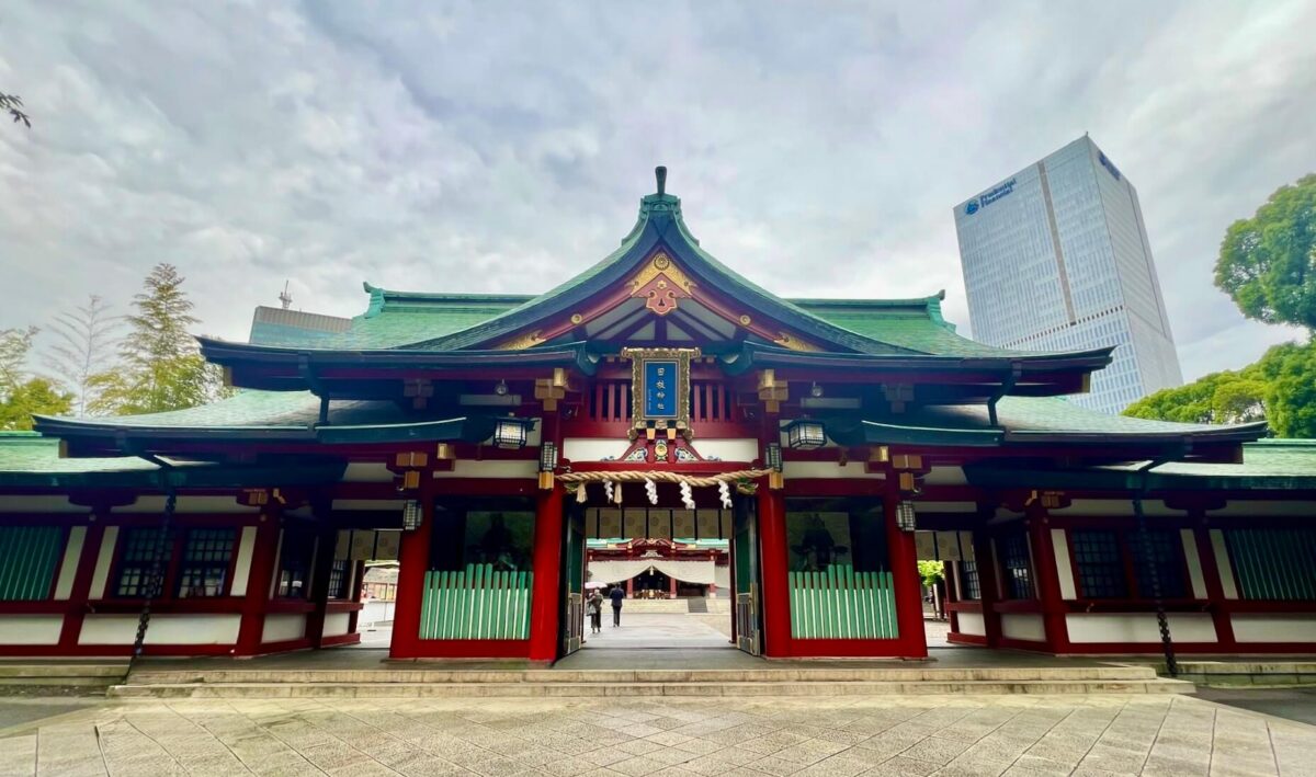 A traditional Japanese Shinto shrine with a green roof and red accents stands before a cloudy sky. Modern skyscrapers and greenery are visible in the background.
