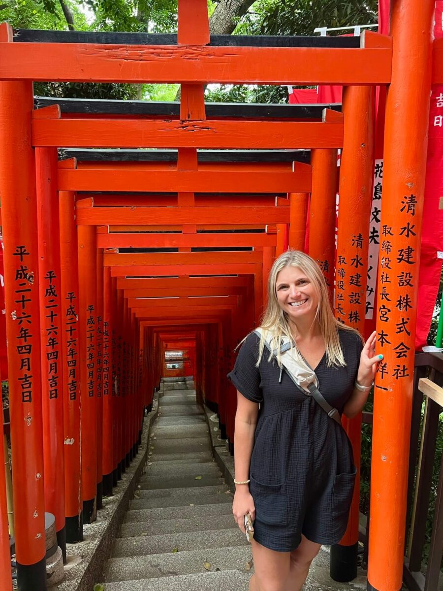 A smiling woman stands beside vibrant red torii gates, with Japanese inscriptions, lining a stone path. The scene exudes a sense of cultural exploration.