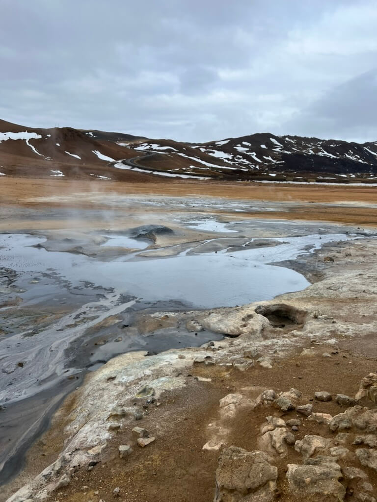 Barren geothermal landscape with steaming hot springs and multi-colored earth, surrounded by snow-dusted hills under a cloudy sky, conveying a desolate yet fascinating atmosphere.