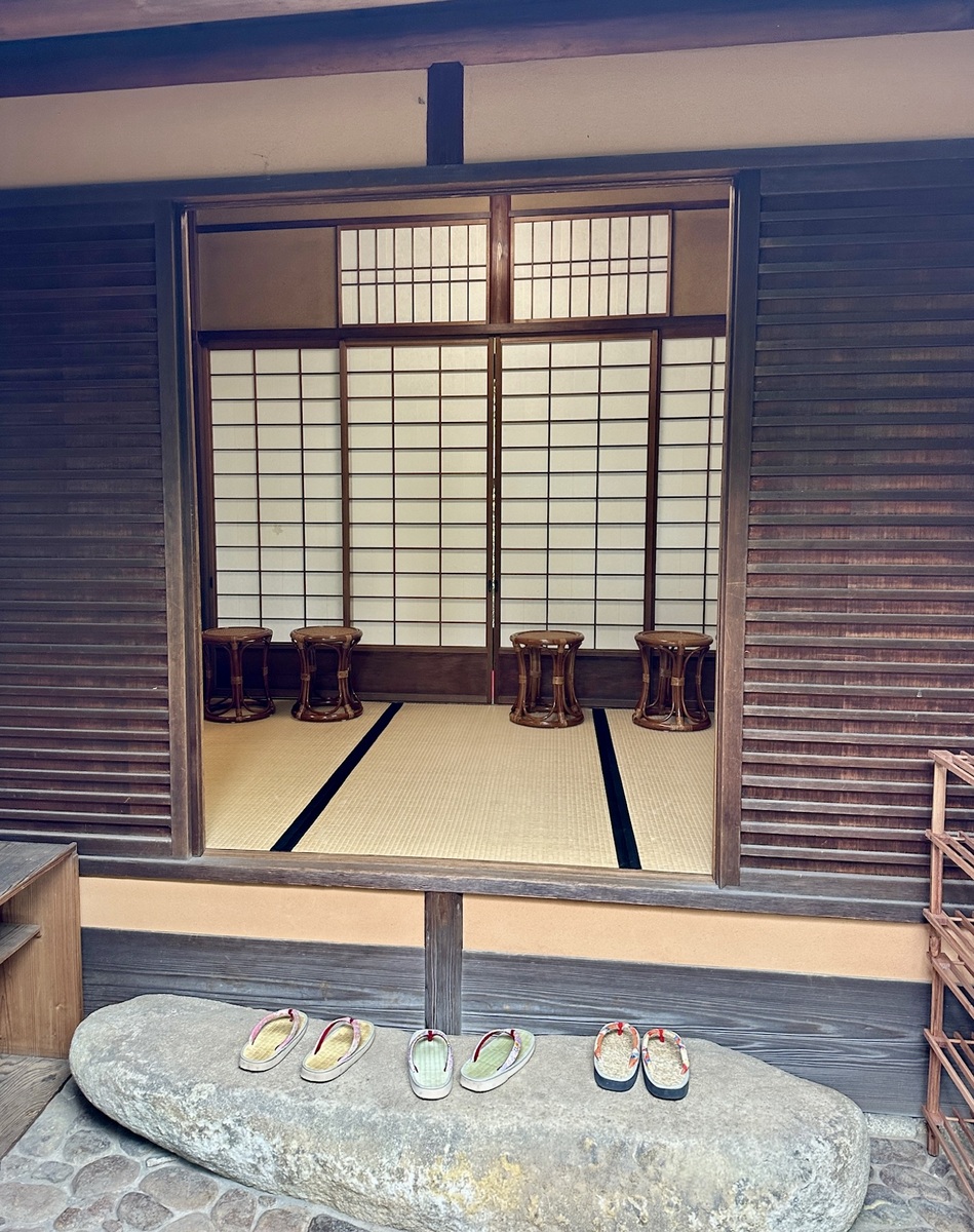 Traditional Japanese room with tatami mats and low wooden stools, viewed through open sliding shoji doors. Three pairs of sandals rest on a stone step.
