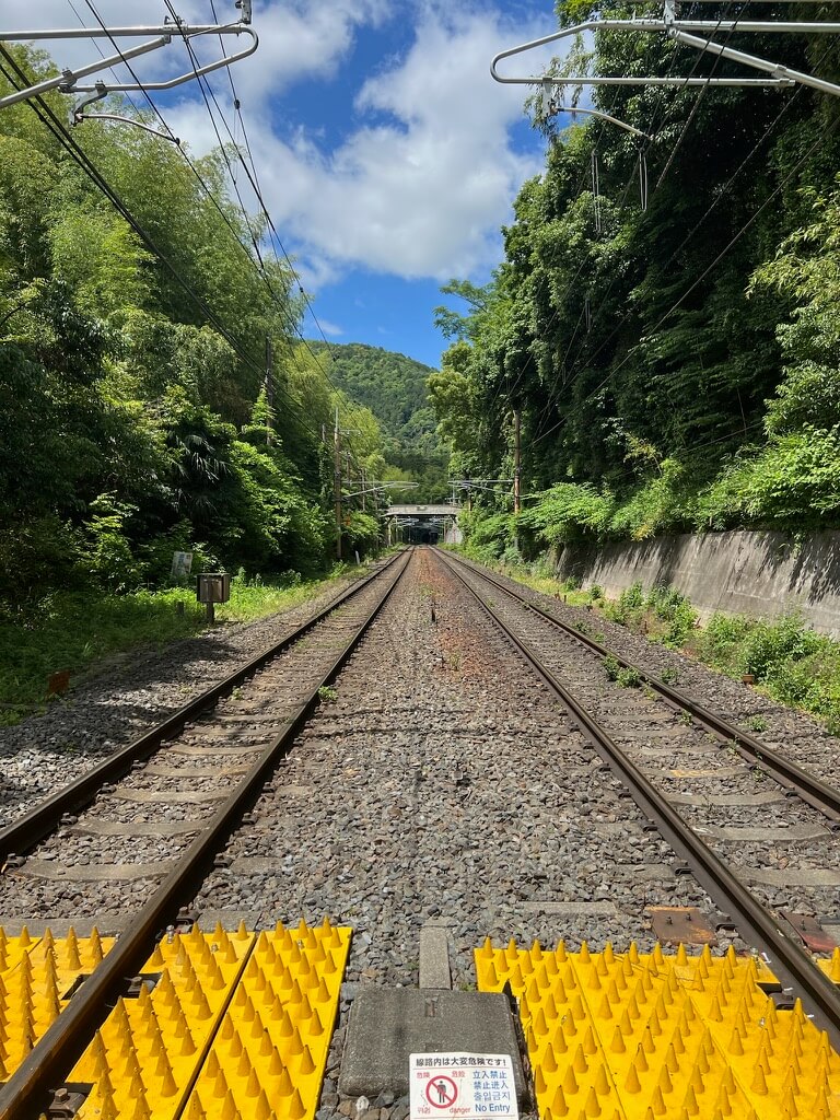 Railway tracks stretch into the distance, flanked by lush green trees under a blue sky. Bright yellow caution spikes and a "No Entry" sign are visible in the foreground.