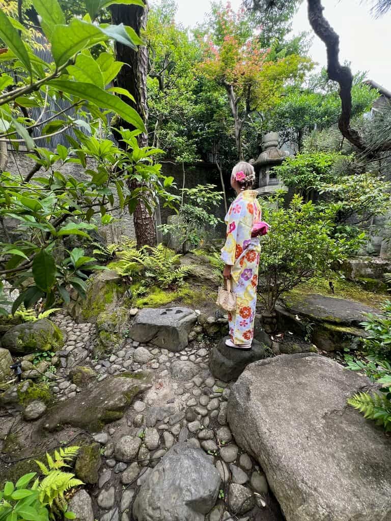 A woman in a colorful kimono with floral patterns stands in a tranquil Japanese garden with lush greenery and stones, conveying serenity and tradition.