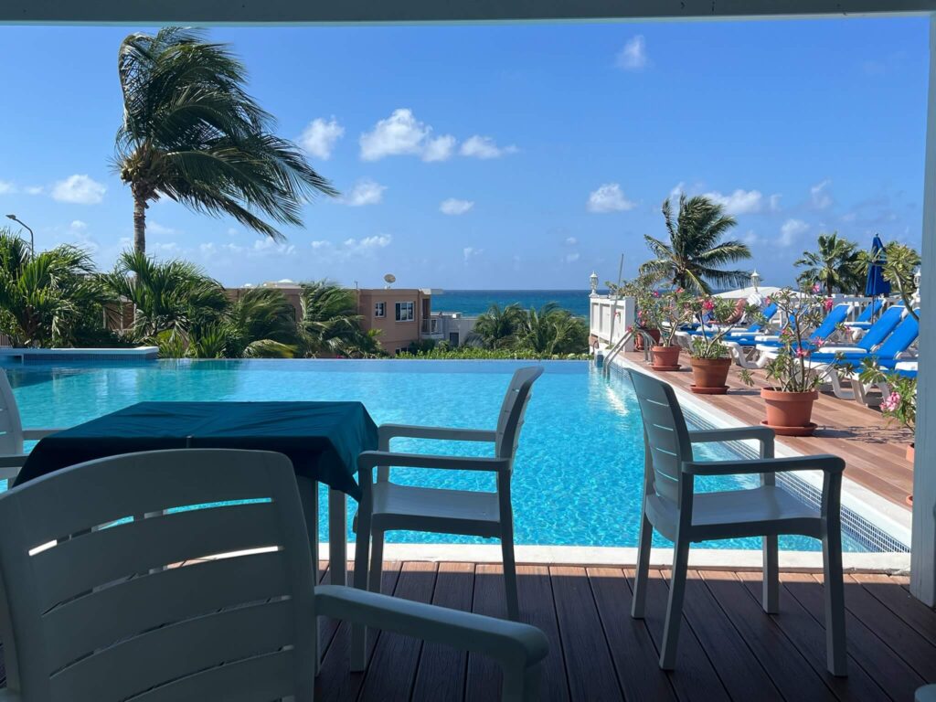 Poolside view with white chairs around a table overlooking a bright blue infinity pool. Palm trees sway gently beside sun loungers under a clear sky.