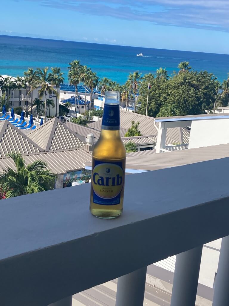 A bottle of Carib Lager sits on a balcony railing overlooking a tropical beach view, with clear blue ocean, palm trees, and a calm, sunny atmosphere.
