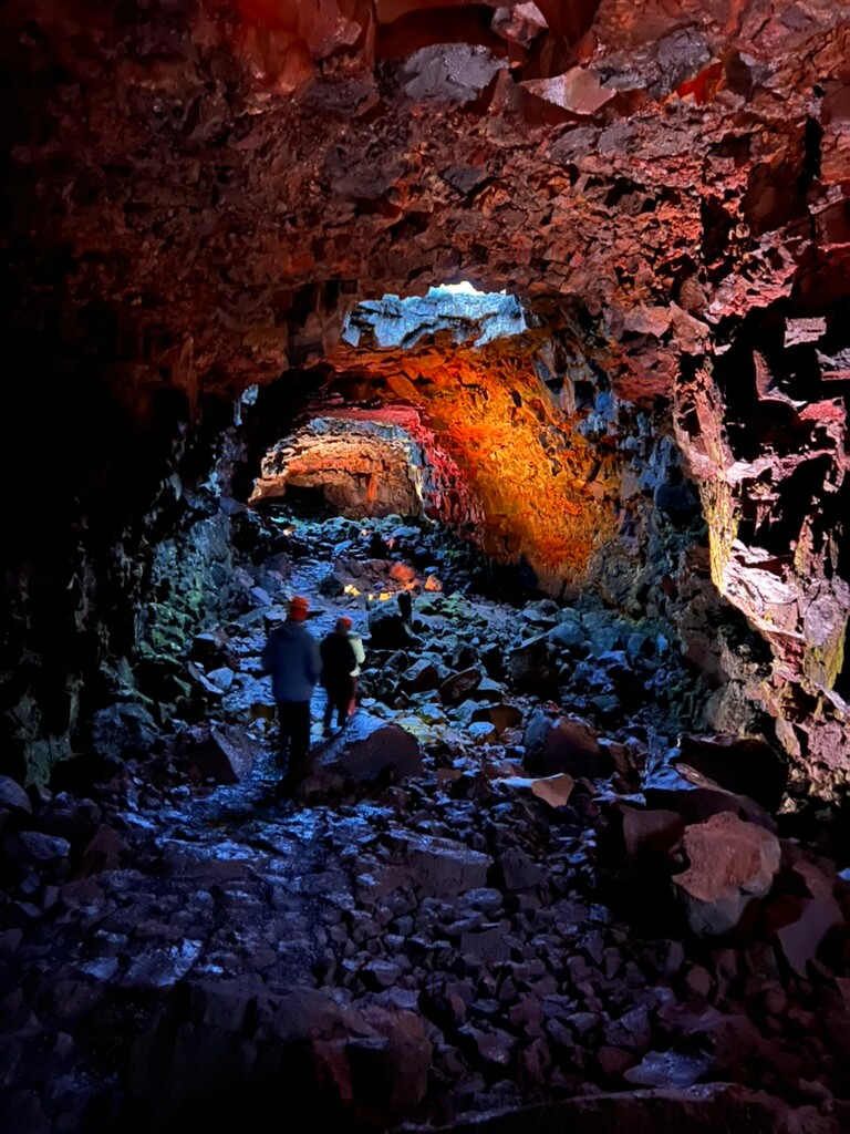 Two people walk through a rocky cave lit by colorful lights, with red, orange, and blue hues highlighting the rough walls and floor, creating a mystical ambiance.