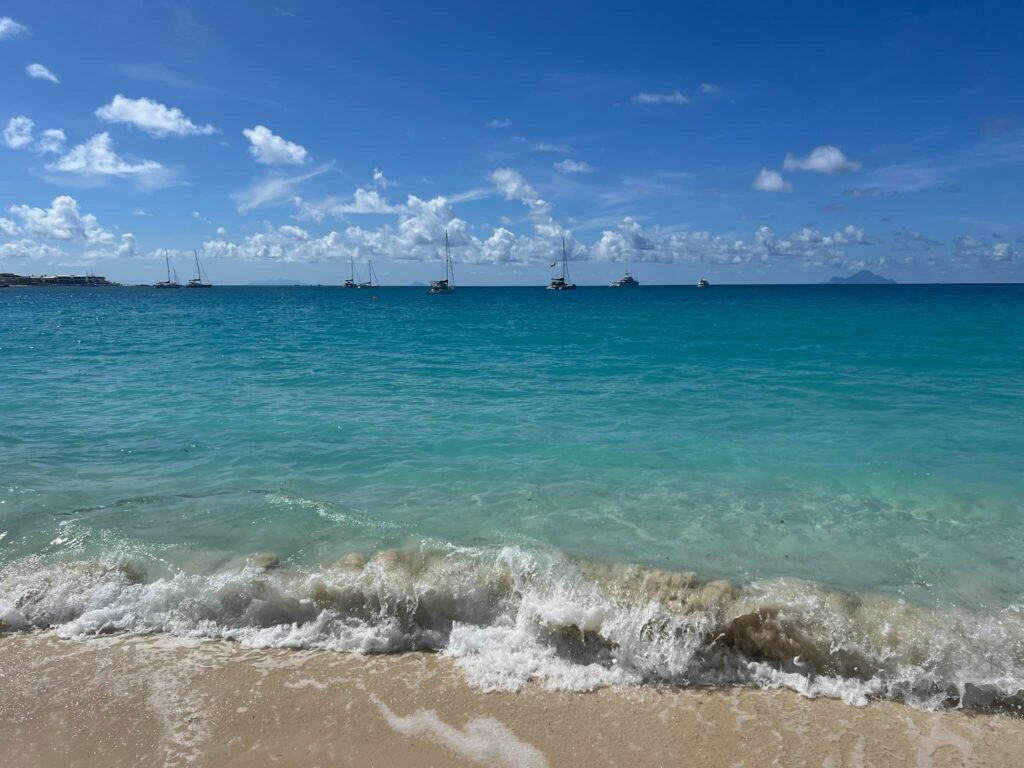 Calm beach scene with gentle waves washing onto sandy shore. Turquoise sea stretches to horizon under clear blue sky, dotted with distant sailboats.