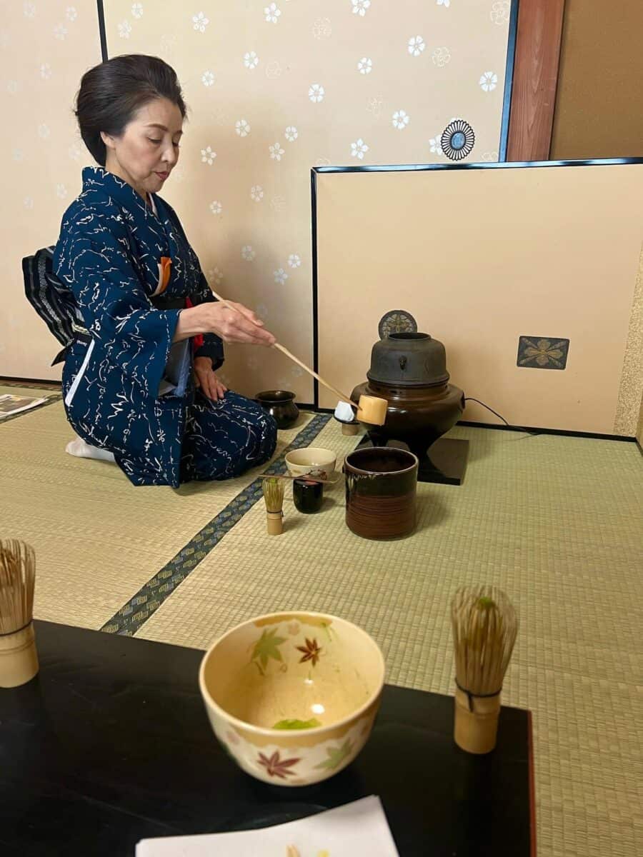A woman in a blue kimono performs a traditional Japanese tea ceremony on tatami mats. She gracefully pours tea, creating a serene and focused atmosphere.