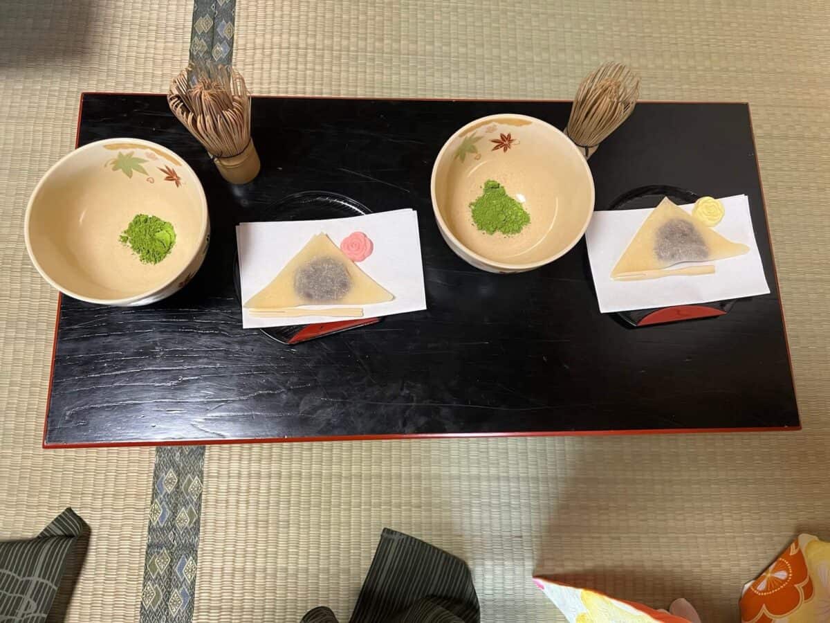 A traditional Japanese tea set with bowls containing green matcha powder alongside wagashi sweets on black trays, set on a tatami mat.