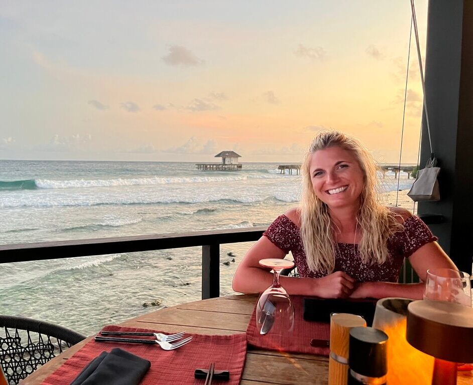 Smiling woman sits at a beachside table during sunset, with waves and an ocean pier in the background. The scene conveys relaxation and warmth.