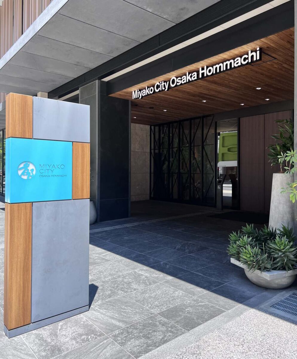 Entrance of Miyako City Osaka Hommachi hotel with a modern design. Features a stylish sign and lush greenery, conveying a welcoming and elegant atmosphere.
