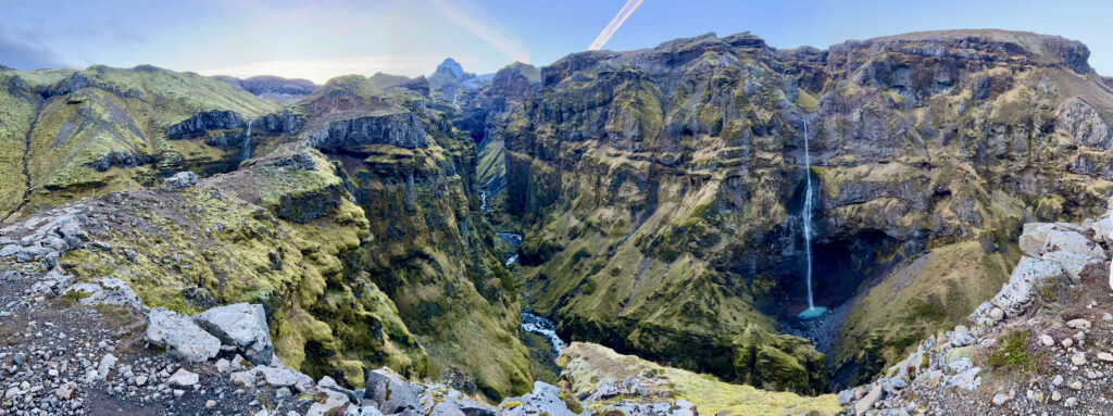 A deep, narrow canyon with steep, moss-covered rocky walls and a small stream running through it. The distant background features rugged mountain peaks under a pale sky. The scene conveys a sense of remote wilderness and natural majesty.