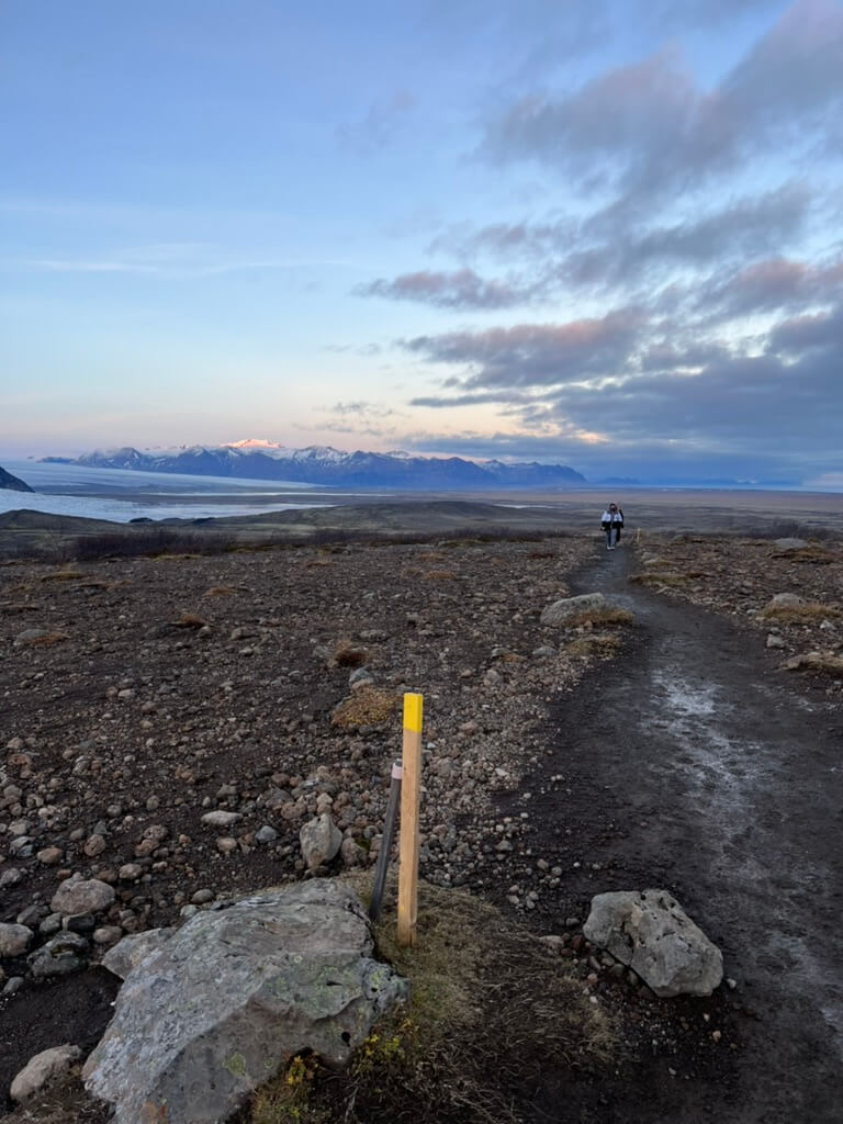 Rocky landscape with a dirt path and distant snowy mountains under a vast, cloudy sky. Two people are walking along the path, creating a serene atmosphere.