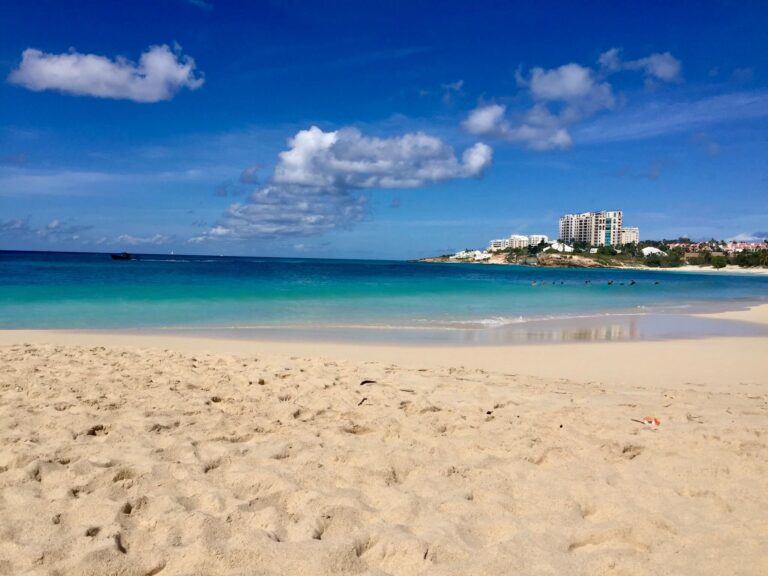 Sandy beach meets turquoise ocean under a blue sky with scattered clouds. A distant shoreline shows buildings, conveying a serene, tropical paradise.