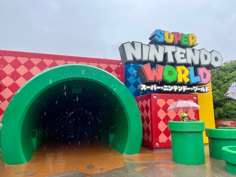 Entrance to Super Nintendo World on a rainy day. Large green pipe foreground, vibrant red walls, and colorful logo. Playful atmosphere.