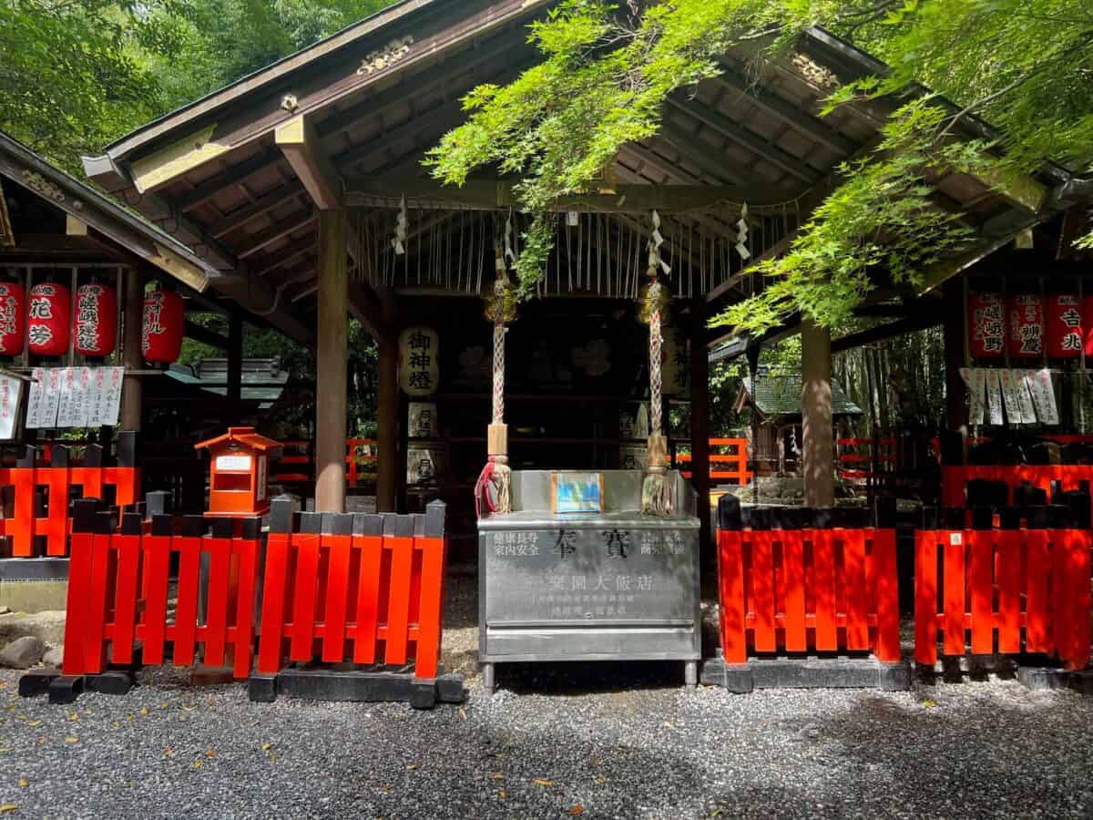 Traditional Japanese shrine with vibrant red and black fencing, paper lanterns, and leafy trees. Sunlight casts patterns on the gravel path, evoking serenity.