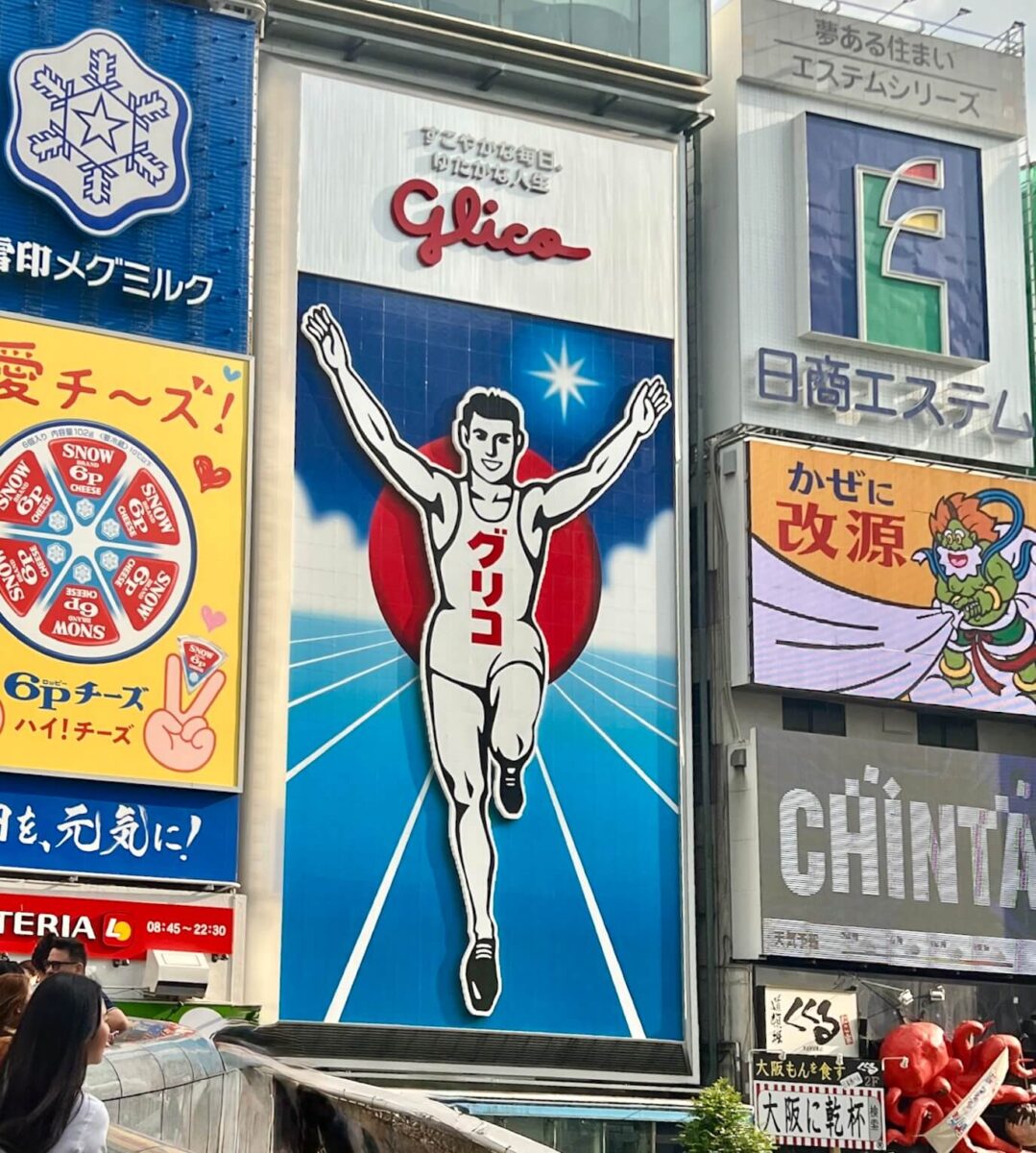 Iconic Glico running man billboard in Osaka, Japan, with arms raised in victory. Bright colors and surrounding commercial advertisements visible. Energetic atmosphere.