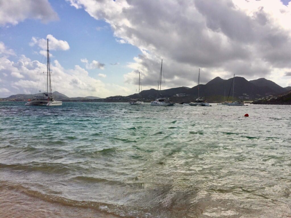Calm seascape with sailboats on turquoise water, under a partly cloudy sky. Mountains in the background add depth, creating a serene atmosphere.
