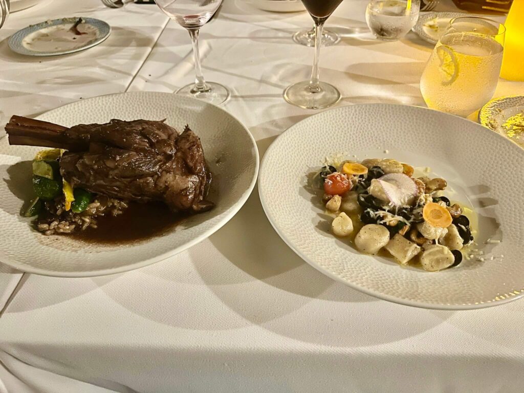 A formal dining setting with two elegant dishes: a lamb shank with vegetables on the left, and creamy gnocchi with vegetables on the right. Glasses and warm lighting convey a sophisticated ambiance.
