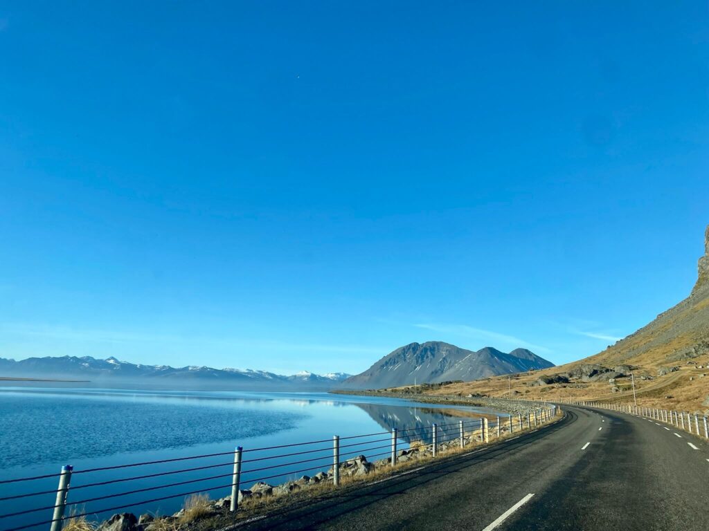 A winding road curves beside a serene blue lake, with snow-capped mountains in the distance under a clear sky, conveying a tranquil and expansive scene.