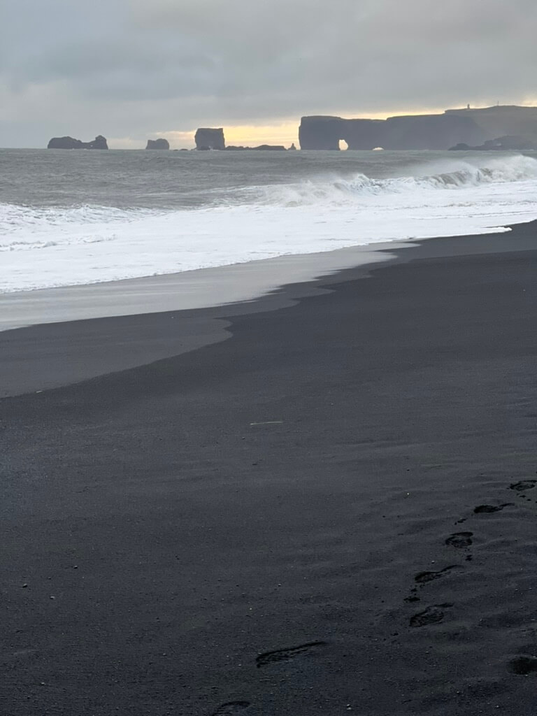 A serene beach scene with dark sand in the foreground and gentle waves approaching. Distant cliffs and rock formations are silhouetted against a cloudy sky.