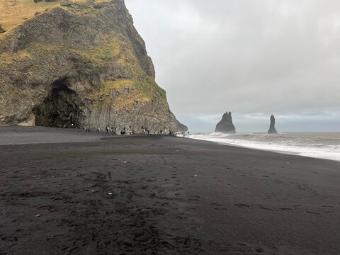 Dramatic black sand beach with towering basalt columns and sea stacks under a gray sky. People explore the cave, adding a sense of scale.