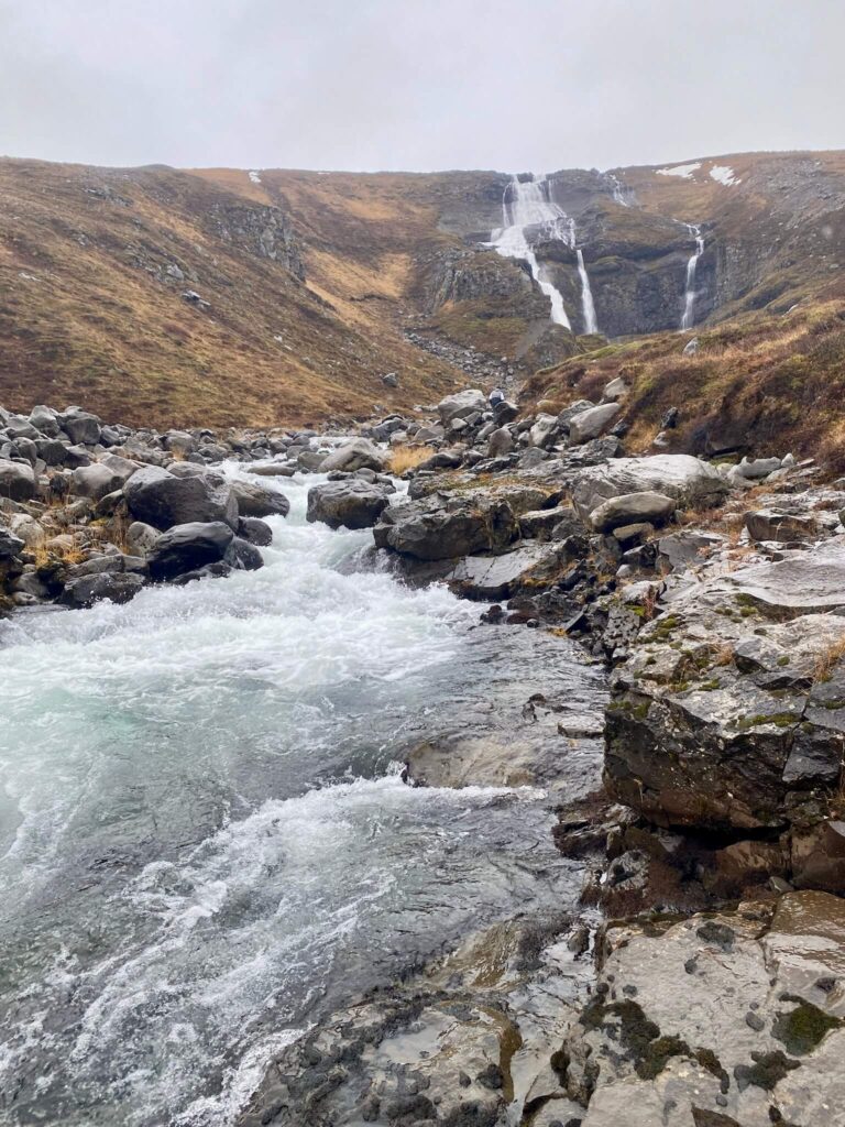 Rocky landscape with a cascading waterfall in the distance, flowing into a clear, rushing stream surrounded by rugged terrain under an overcast sky.