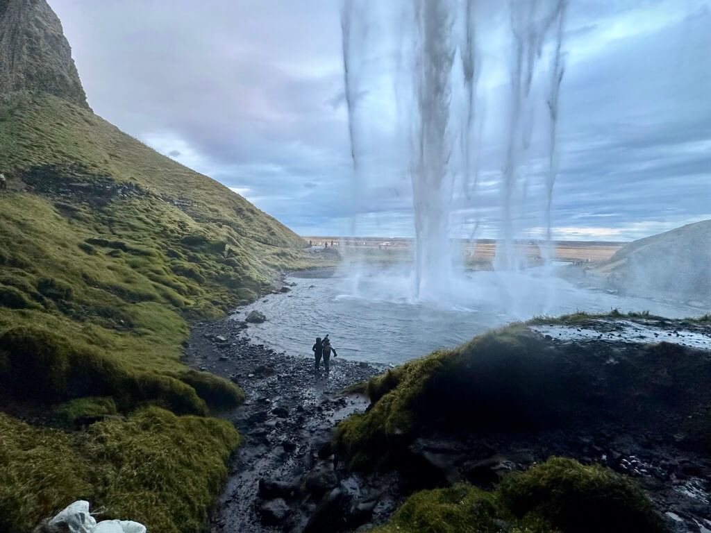 View from behind a waterfall in a lush, green landscape. Two people in the distance admire the scene under a gray, cloudy sky, conveying tranquility and awe.