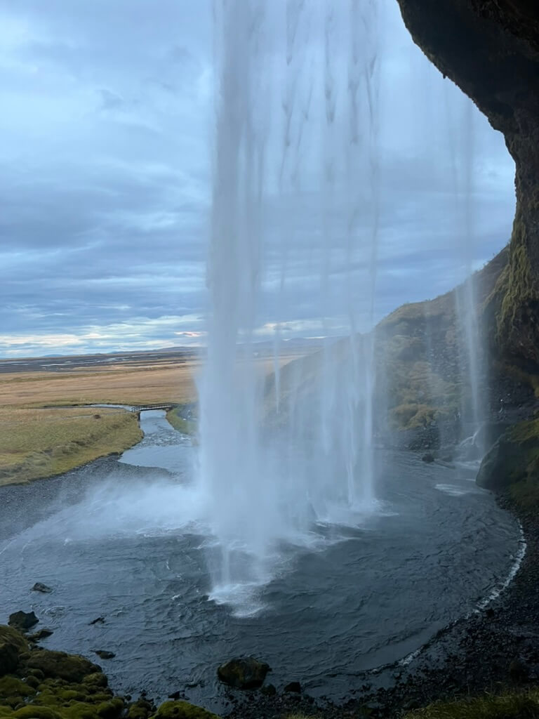 View from behind a tall waterfall, cascading into a pool surrounded by mossy rocks and open fields. The sky is overcast, creating a serene atmosphere.