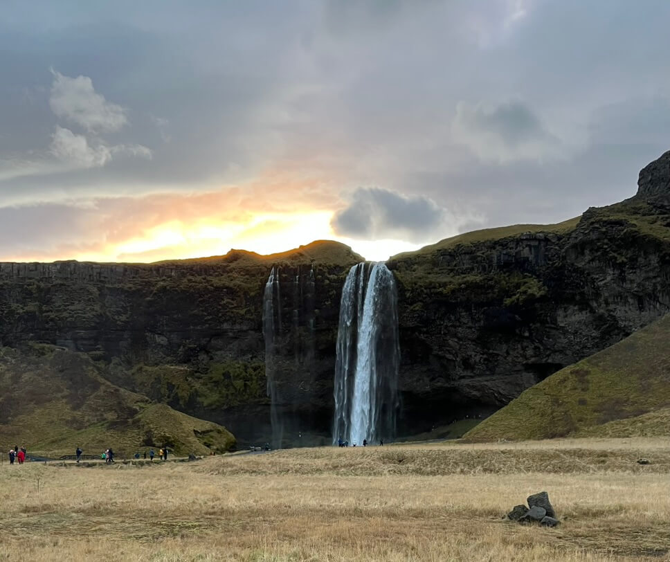 A majestic waterfall cascades over a mossy cliff at sunset. The sky glows warmly, and a few people are scattered on the grassy field below.