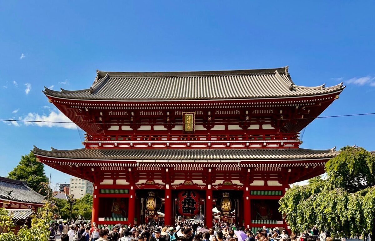 Vibrant red and white traditional Japanese temple gate under a clear blue sky, crowded with visitors, conveying a lively, bustling atmosphere.