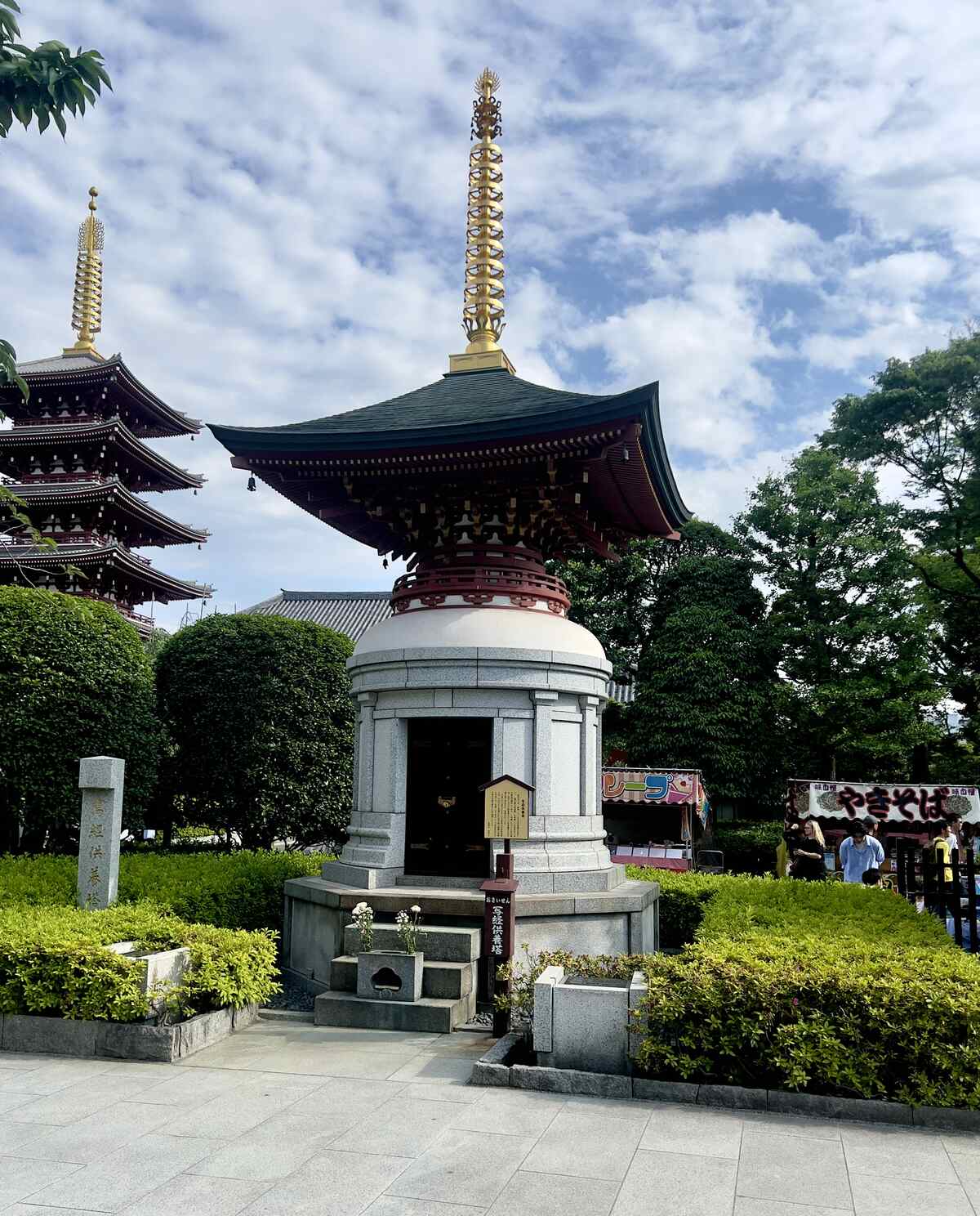A serene scene depicts a Japanese temple courtyard with a pagoda. Lush greenery surrounds the ornate structures under a partly cloudy sky. Visitors in the background.