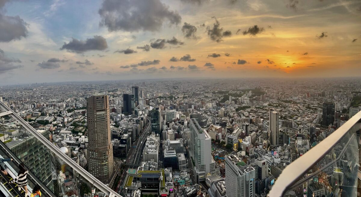 Aerial view of a bustling cityscape at sunset, high-rise buildings visible under a vibrant sky with scattered clouds, casting a serene, warm glow.