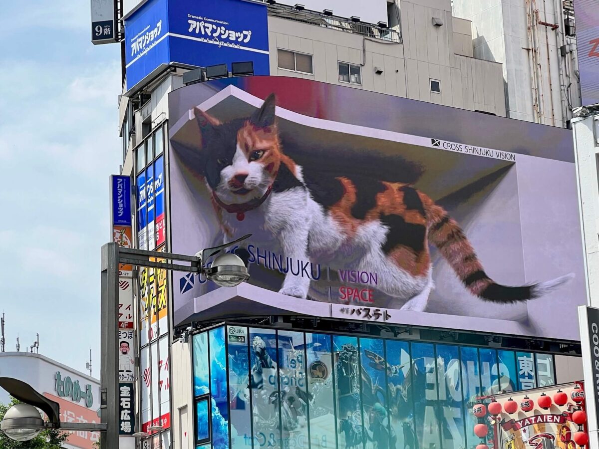 A large 3D billboard in Shinjuku features a lifelike calico cat playfully emerging. The vibrant urban setting includes Japanese signage and a cloudy sky.