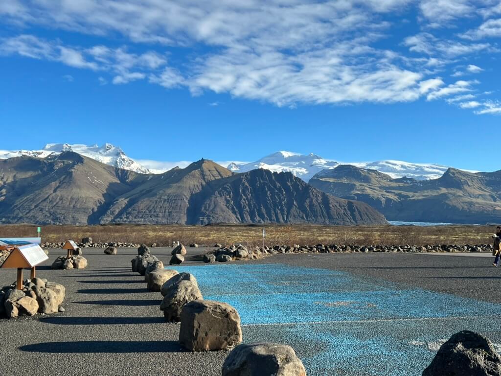 Rugged snow-capped mountains under a clear blue sky dominate the background, with a rocky parking area and blue-lined parking spot in the foreground.