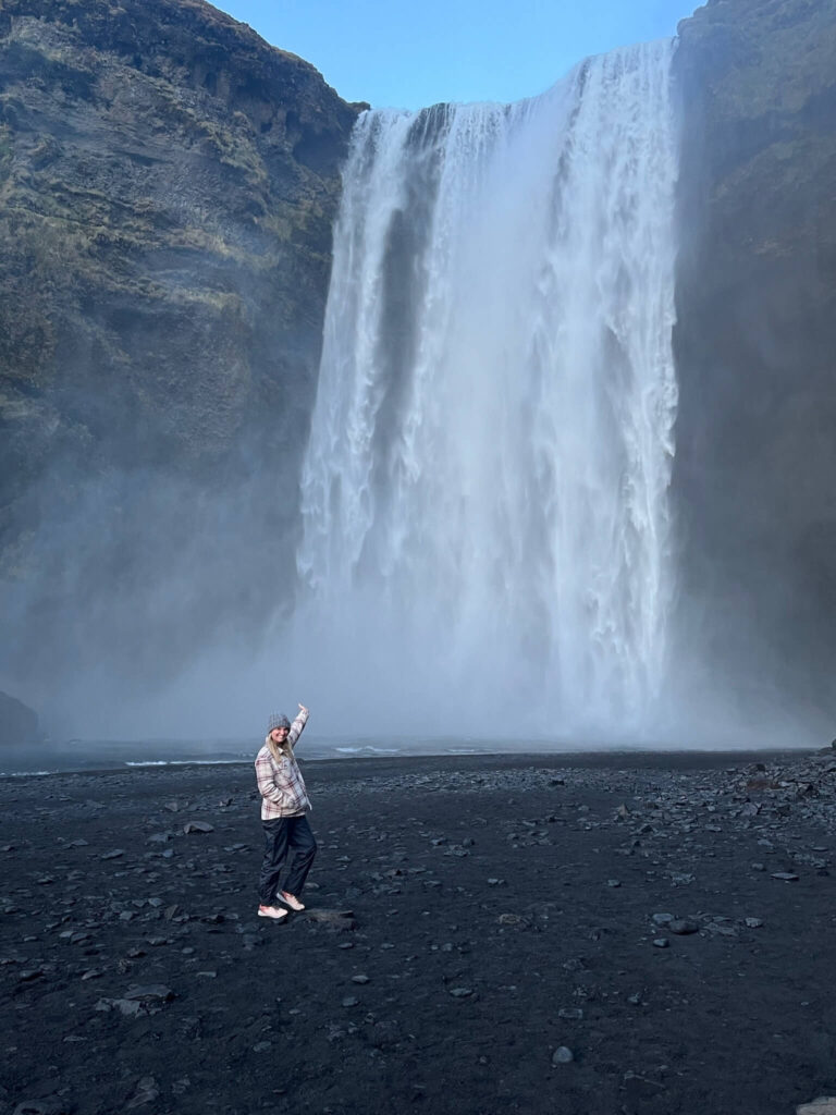 A person stands joyfully on a rocky, dark terrain, pointing up at a towering, misty waterfall cascading down a steep cliff under a clear blue sky.