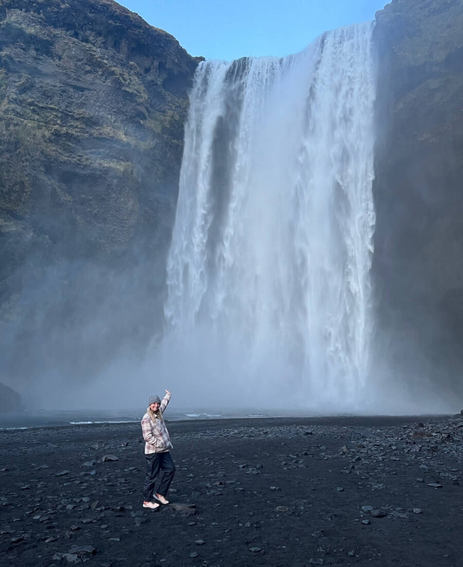 A person stands joyfully on a rocky, dark terrain, pointing up at a towering, misty waterfall cascading down a steep cliff under a clear blue sky.